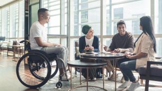 Quatre personnes issues de cultures diverses, dont un homme en fauteuil roulant, échangent dans un espace lumineux et convivial, favorisant l’inclusion.
